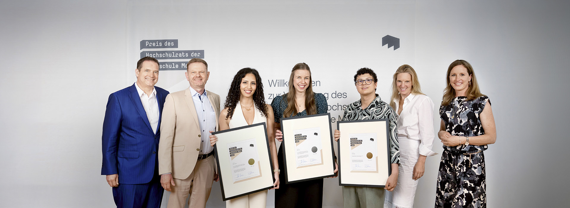 Die Preisträger:innen des Hochschulratspreises 2026 Lina Abu Yousef, Sarah Schütz und Thomas Range gemeinsam mit Hochschulpräsidentin Katharina Dahm und dem Hochschulratsvorsitzenden Michael Maskos vor der Fotowand der Preisverleihung.