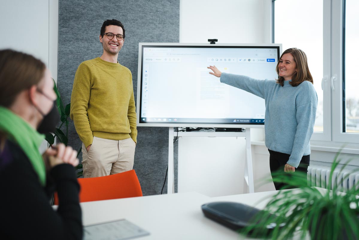 Zwei Personen stehen vor einer digitalen Tafel und eine Person sitzt an einem Tisch.