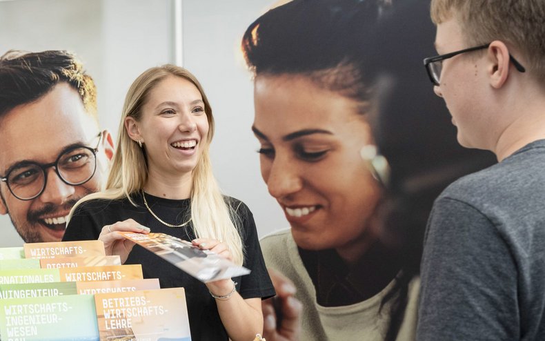 Mitarbeitende der Allgemeinen Studienberatung informieren Studieninteressierte an einem Messestand der Hochschule Mainz über Studienmöglichkeiten und Studiengänge.