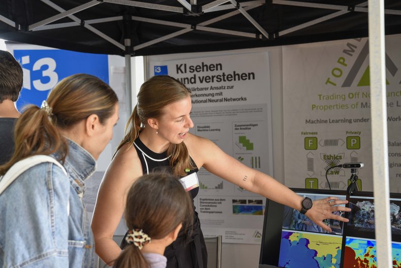 Eine Mitarbeiterin der Hochschule Mainz erklärt Besucherinnen und Besuchern an einem Messestand ein Forschungsprojekt am Mainzer Wissenschaftsmarkt.
