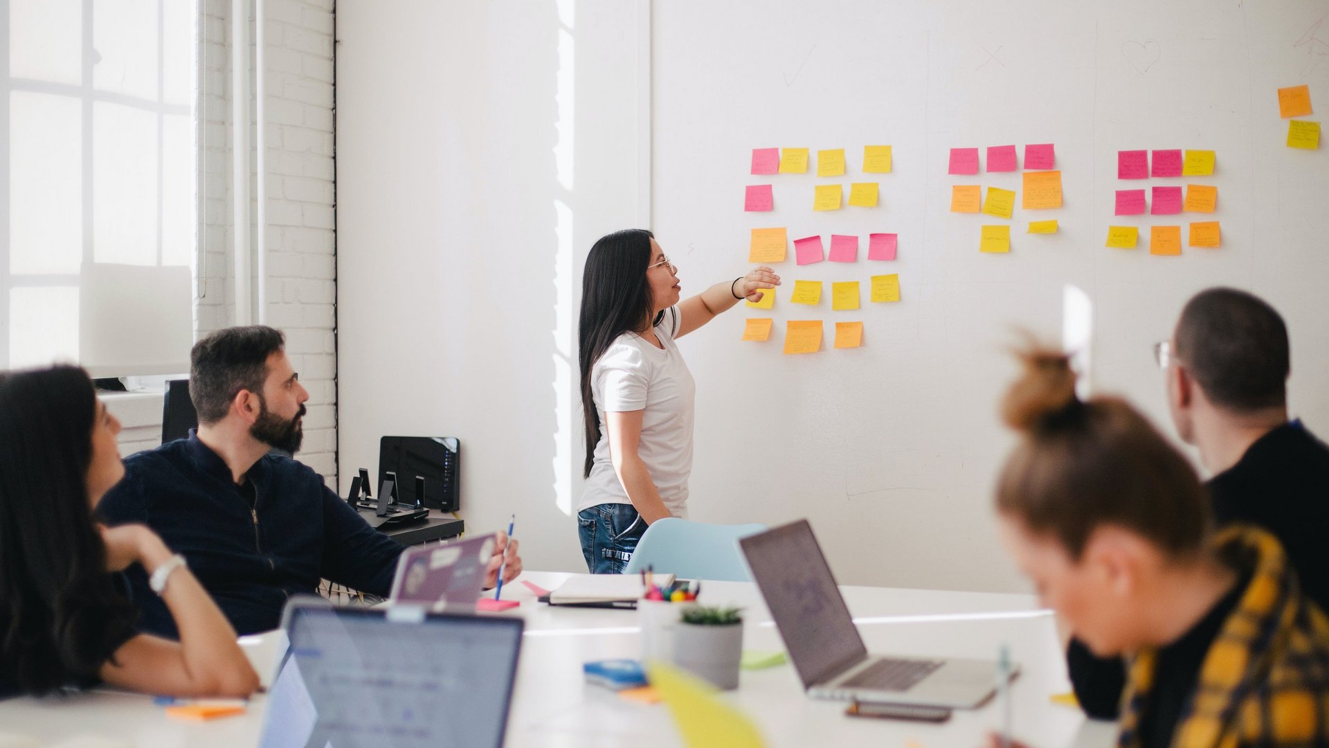 Eine Person präsentiert etwas mit PostIts an einer Wand. Vier Personen sitzen davor an einem Tisch mit Laptops.