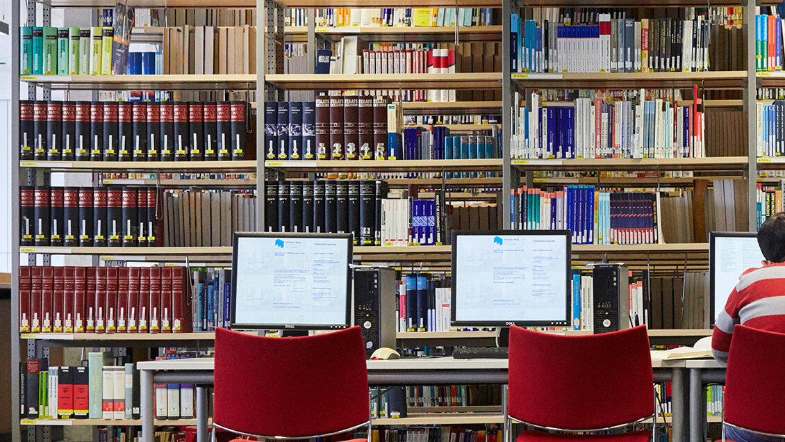 Bücherregal in der Bibliothek, im Vordergrund zwei PCs für die Katalogrecherche